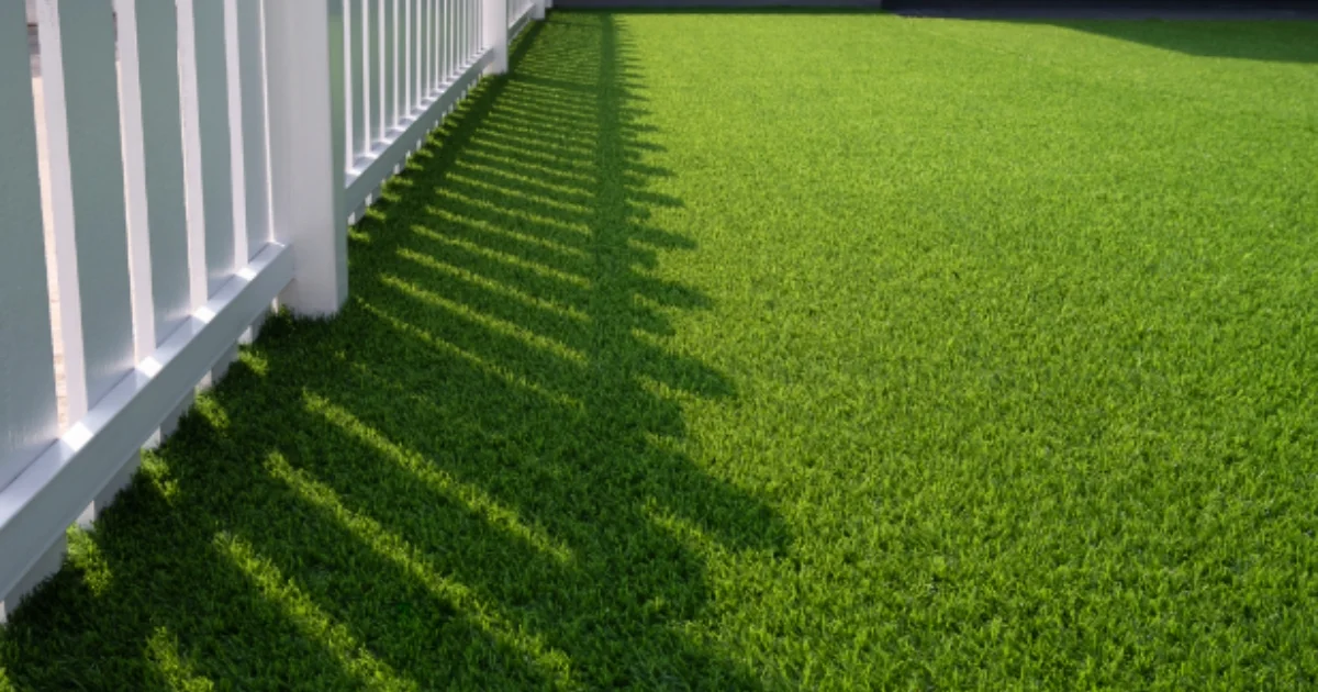 Close-up of artificial grass casting long shadows from a white fence in a backyard or patio setting.